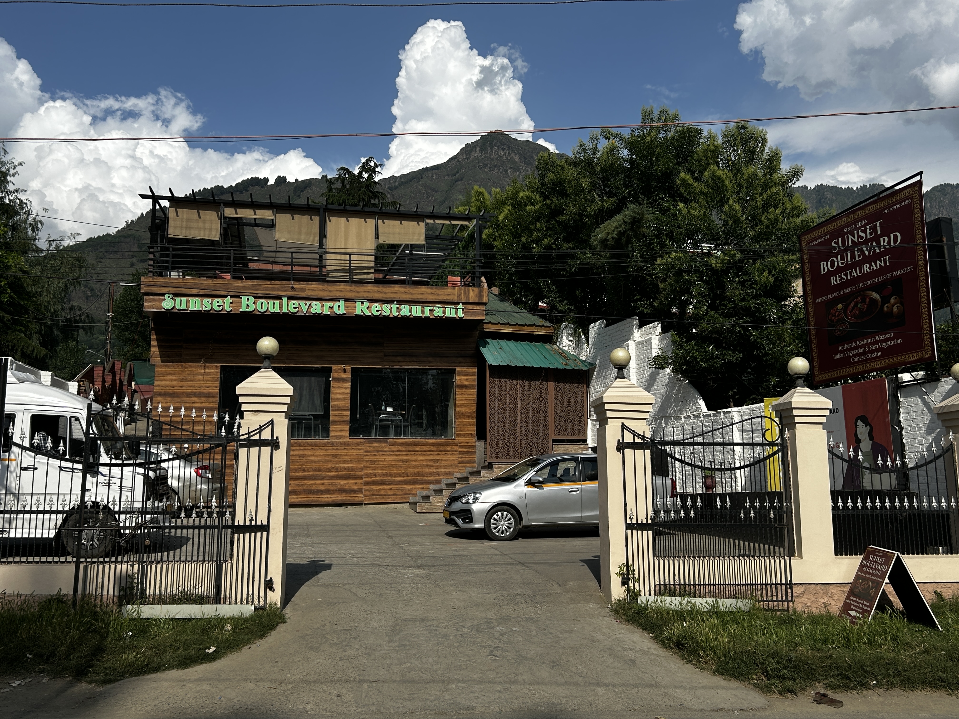 Wide view of Sunset Boulevard Restaurant's entrance - wooden facade, gate, driveway and parking, with mountains in the background